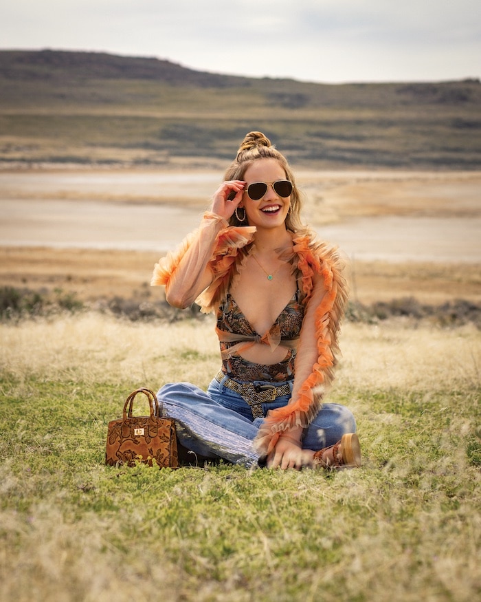 A smiling woman sits on grass in a scenic, open landscape. Stylish in sunglasses and a colorful top with orange sheer sleeves, jeans, and holding a brown purse, she enjoys the outdoors with her solar powered confidence and relaxed vibe.